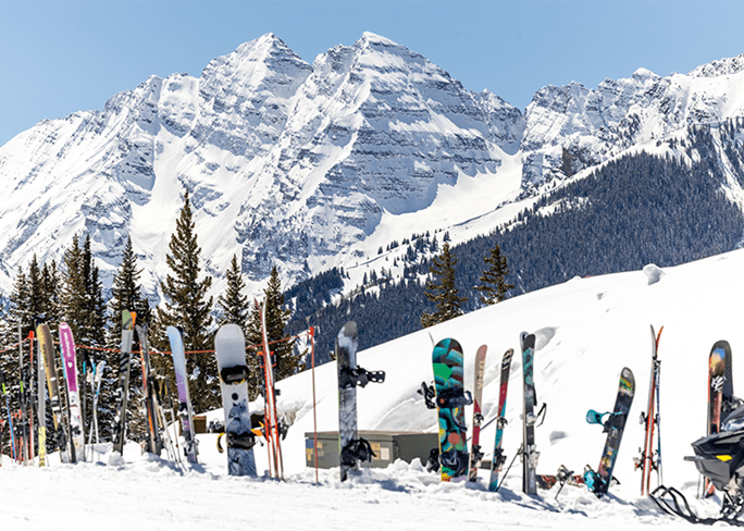 Skies and snowboards lined up in the snow, outside an Aspen Snowmass restaurant, with the maroon bells in the background